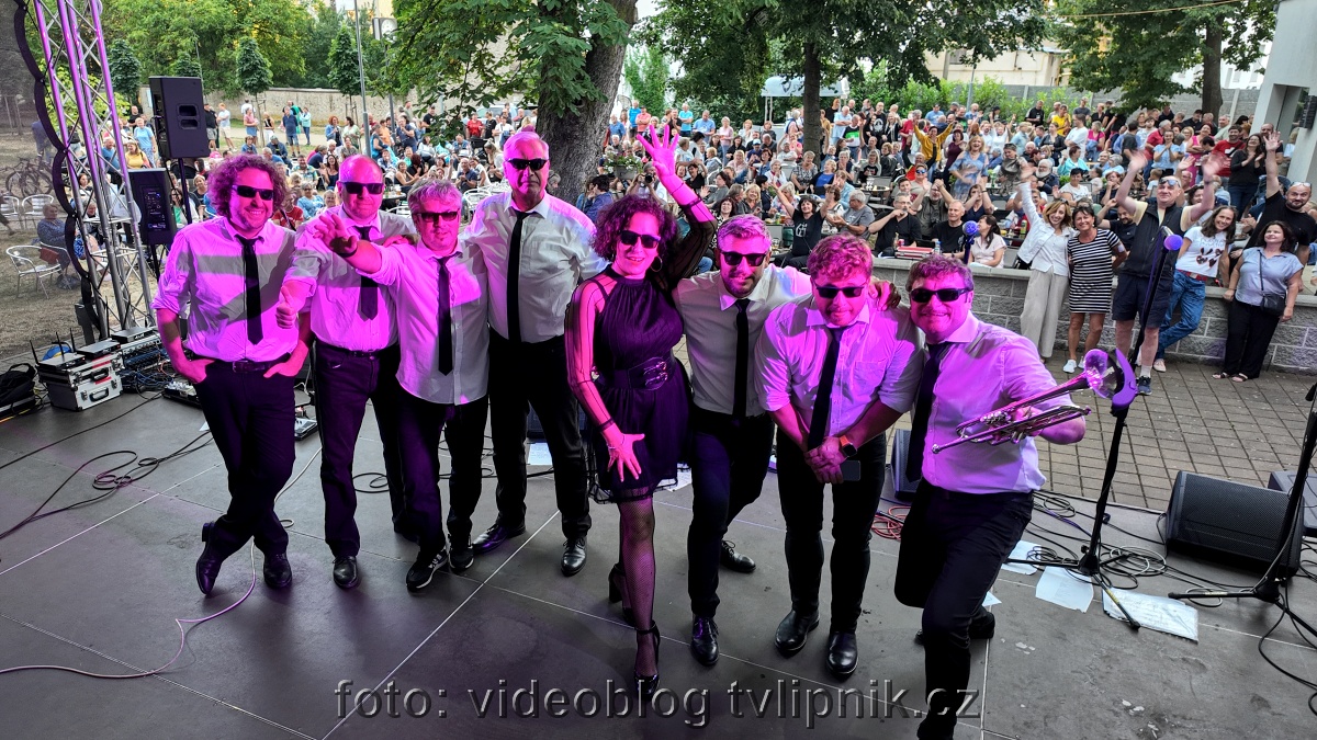 Skupina muzikantů v růžovém světle pózuje před velkým publikem na venkovním koncertě, mávají a hrají.